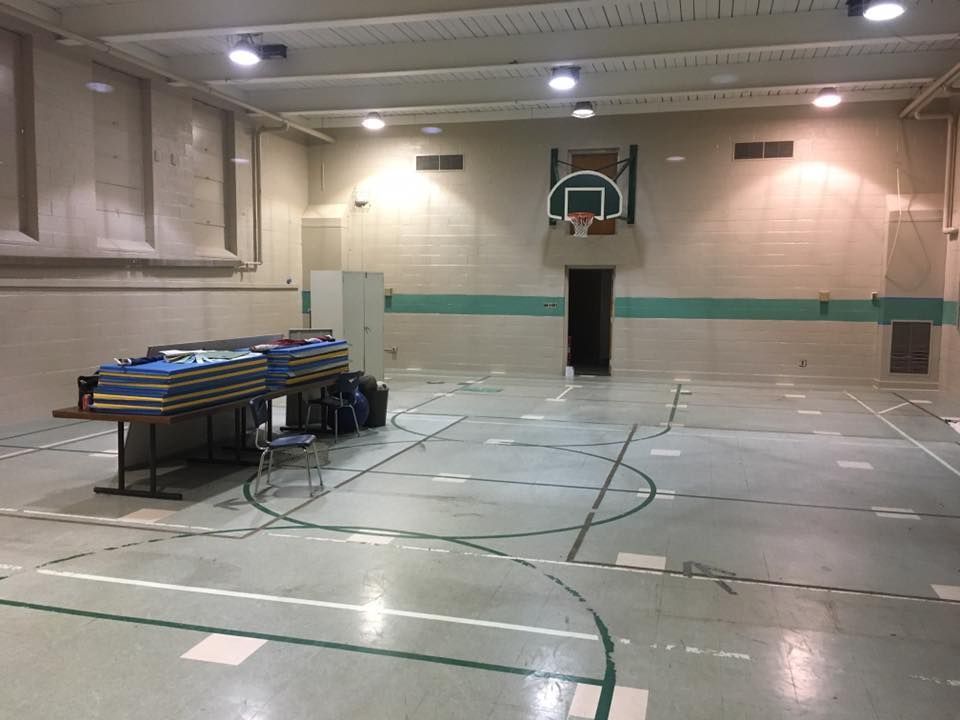 Indoor basketball gym with a hoop and stacked tables on the left, empty polished floor in the foreground