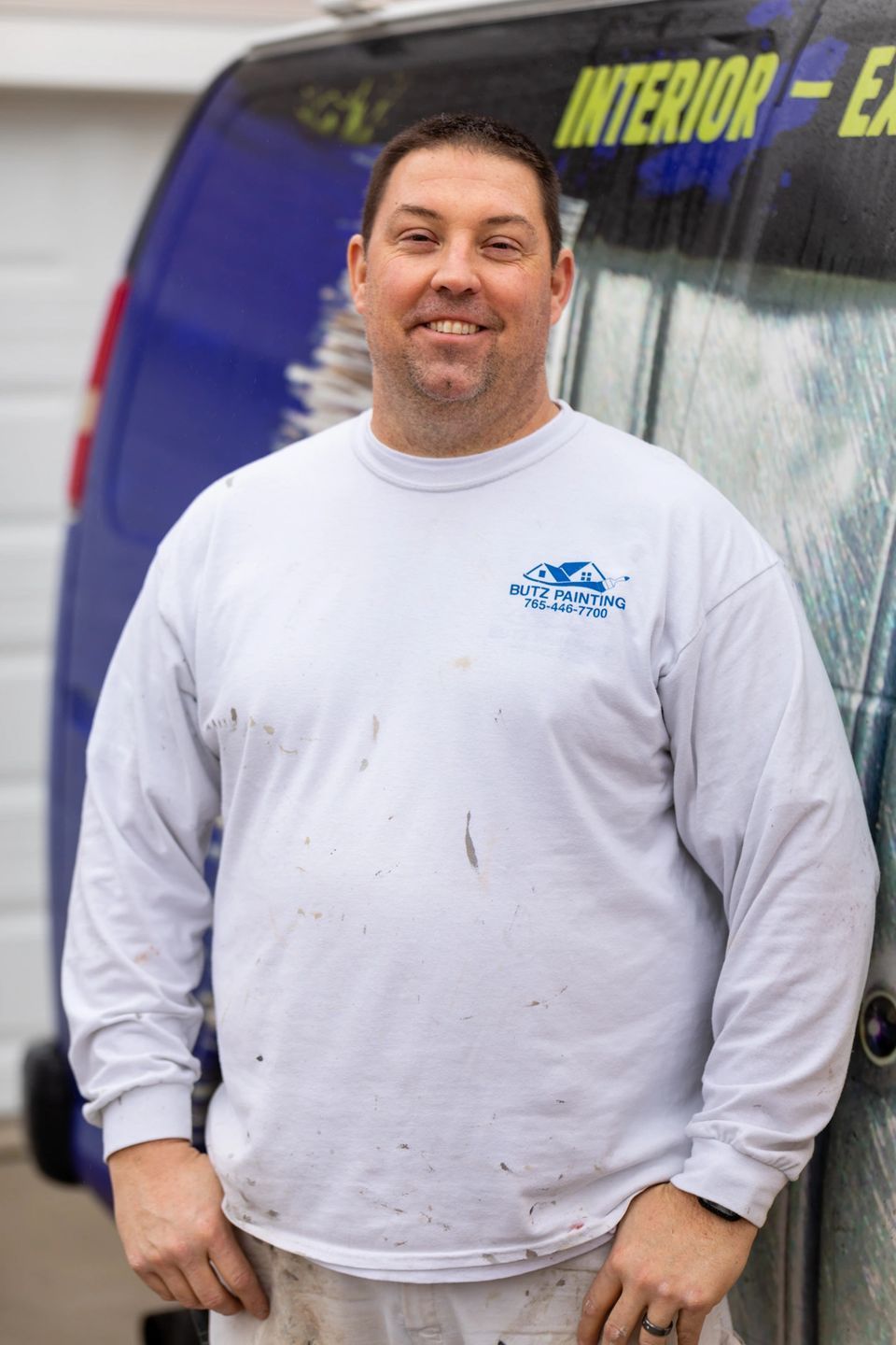 Man standing in front of a vehicle, wearing a white long-sleeve shirt and smiling.