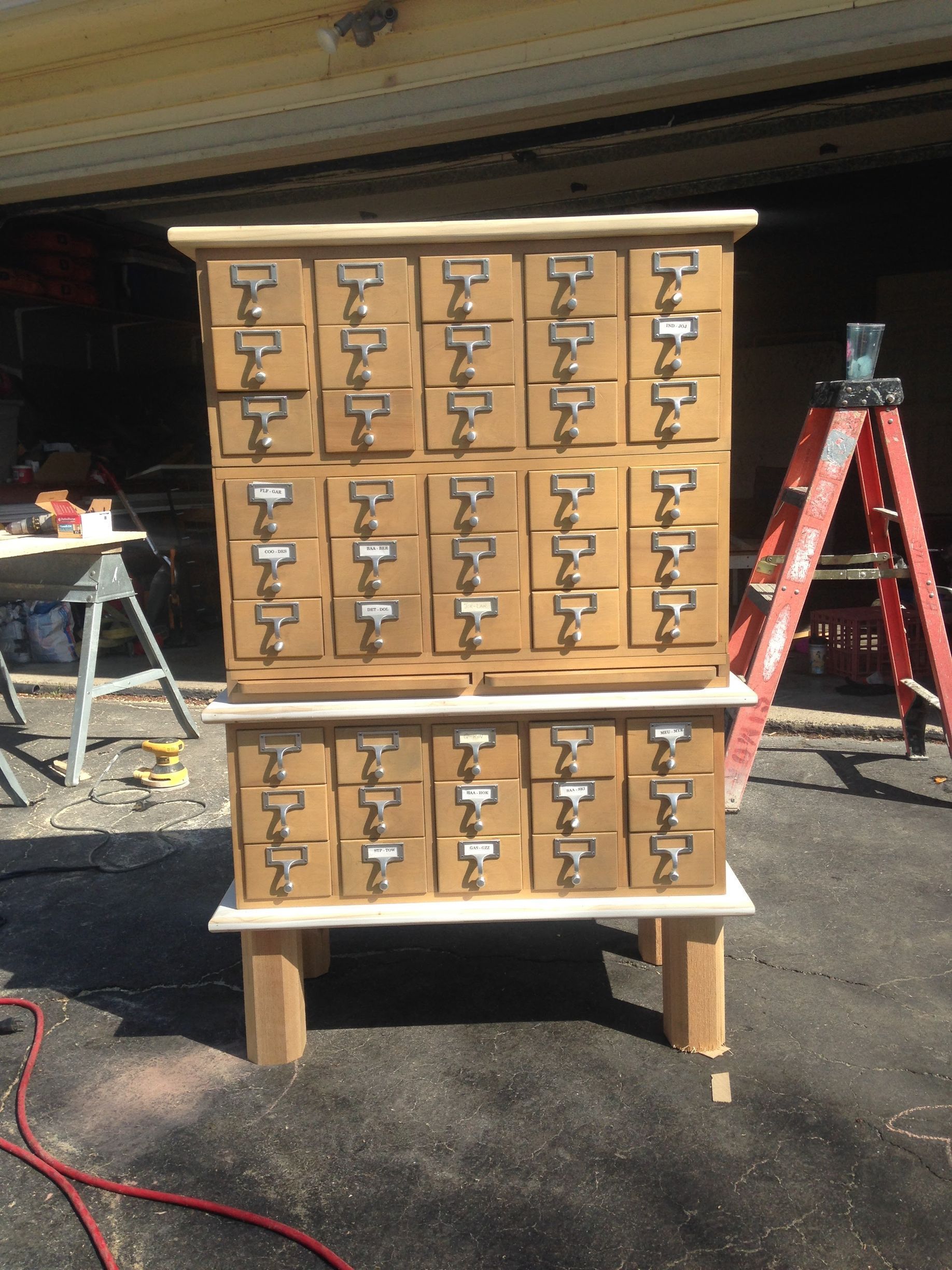 Wooden cabinet with many small drawers and metal handles, standing outdoors on pavement near a ladder.