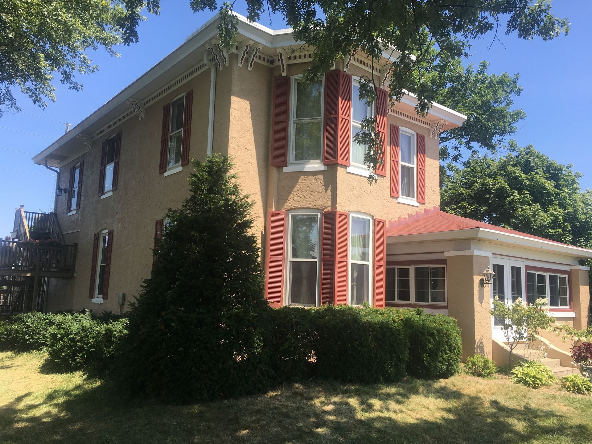 Two-story tan house with red shutters and a small porch under a sunny blue sky