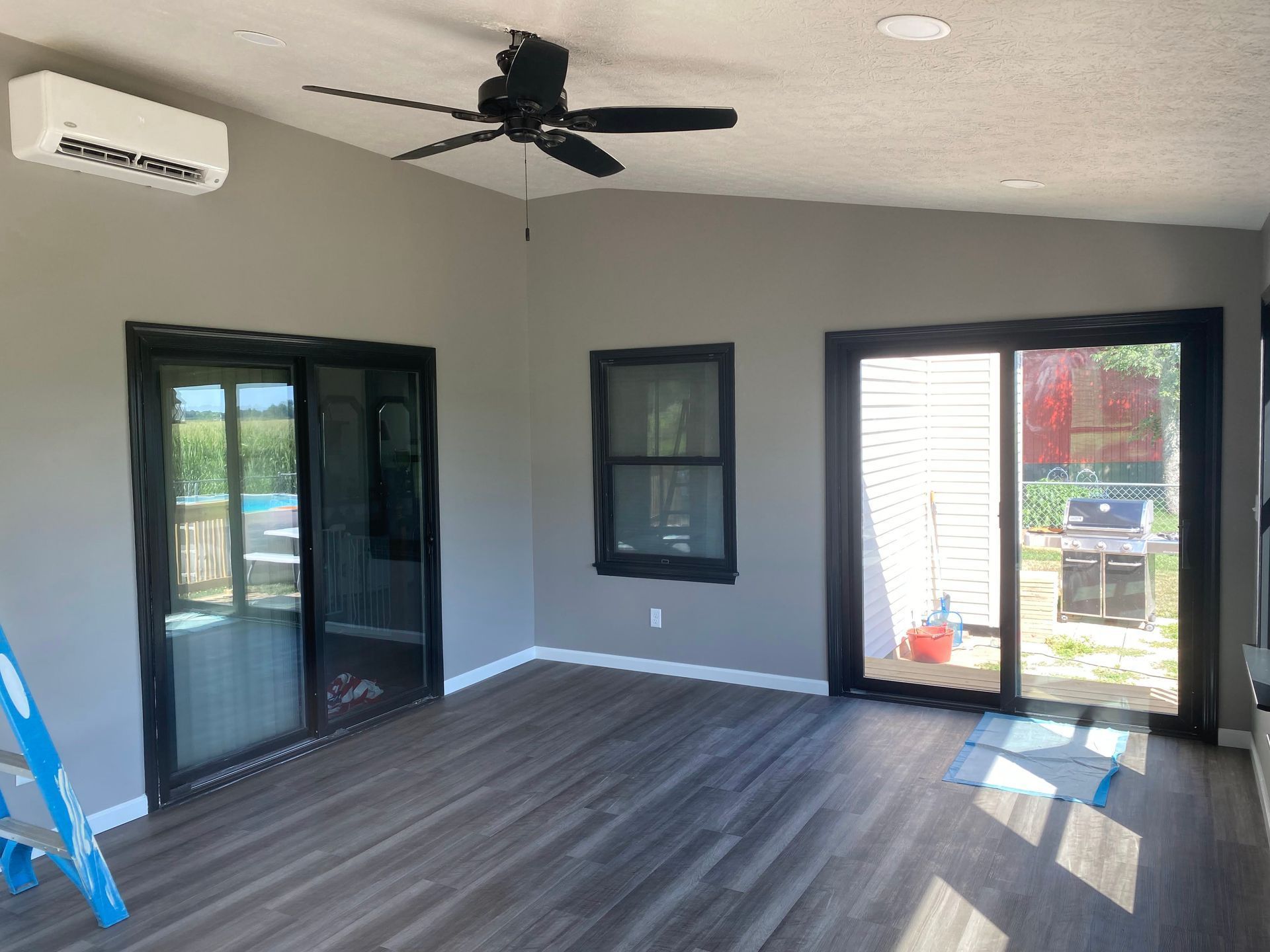 Empty covered patio with gray floors, ceiling fan, sliding glass doors, and a window overlooking a backyard.