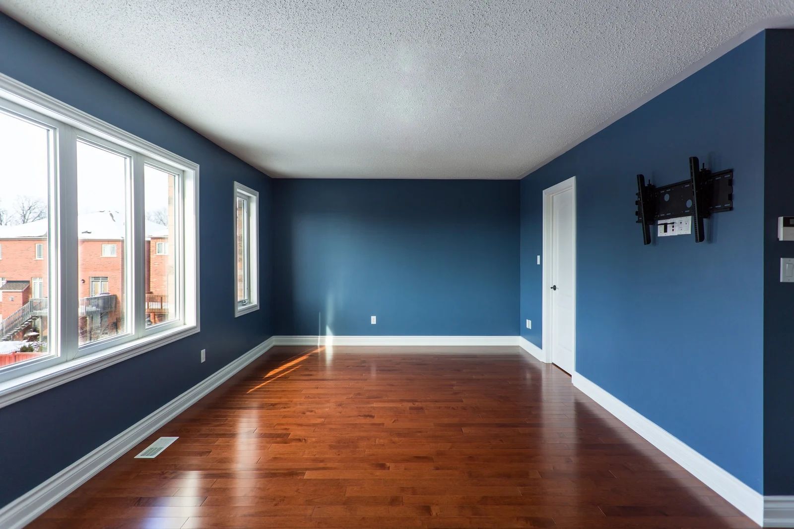 Empty room with blue walls, hardwood floor, white trim, windows, and a mounted TV bracket.