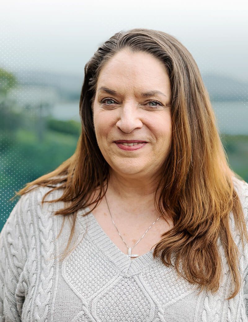 Woman with long brown hair, in a gray cable-knit sweater, smiles outdoors, silver cross necklace.
