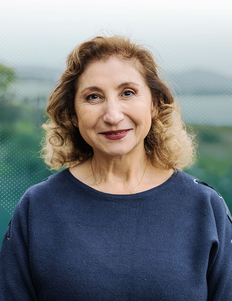 Woman with curly brown hair in blue sweater, smiling, against a blurry landscape backdrop.