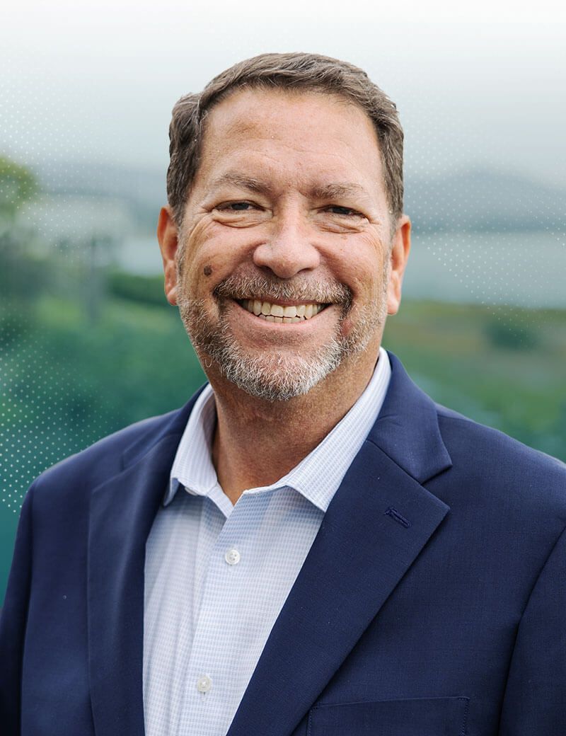 Smiling older man in a navy blazer outdoors with a blurred natural background.