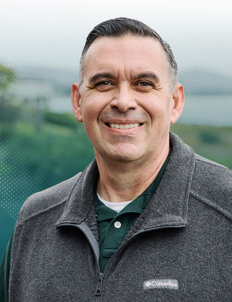 Smiling man in gray fleece jacket, outdoor setting with green and blue background.