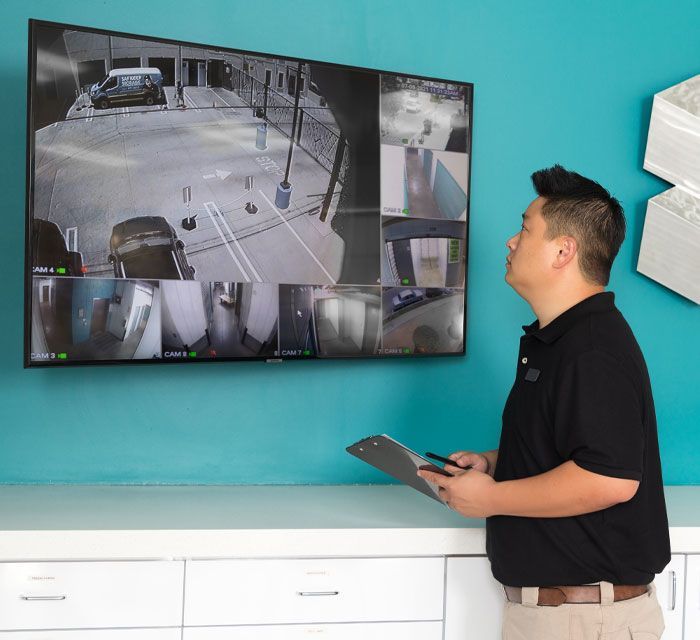 Man in black shirt monitoring security cameras on a large screen in a white room.