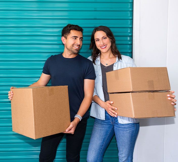 A smiling couple stands in front of a teal storage unit door, holding moving boxes. 