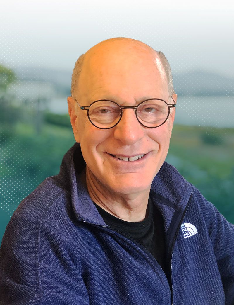 Smiling bald man wearing glasses and a blue jacket, standing outdoors with a blurred background.