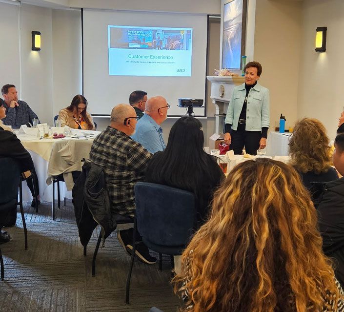 Woman presenting to group at a table. Presentation screen visible. Attendees seated, listening.