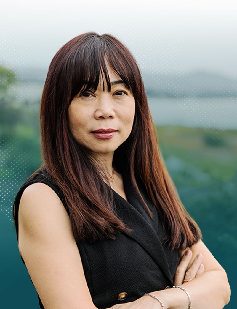 Woman with long dark hair and bangs, arms crossed, wearing a black top. Blurred outdoor background.