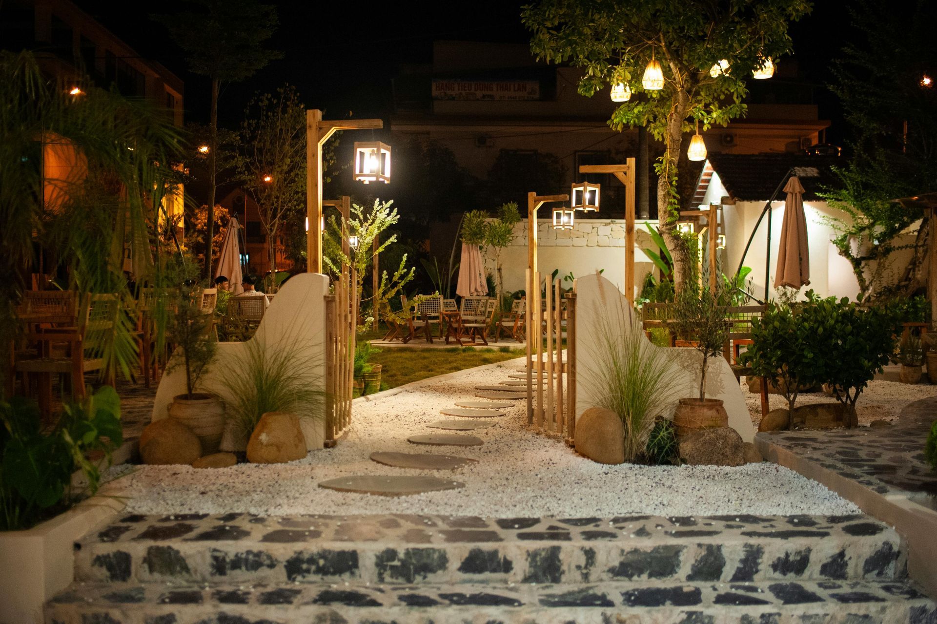 Stone steps lead to a lit garden path at night, framed by lanterns, plants, and white gravel.