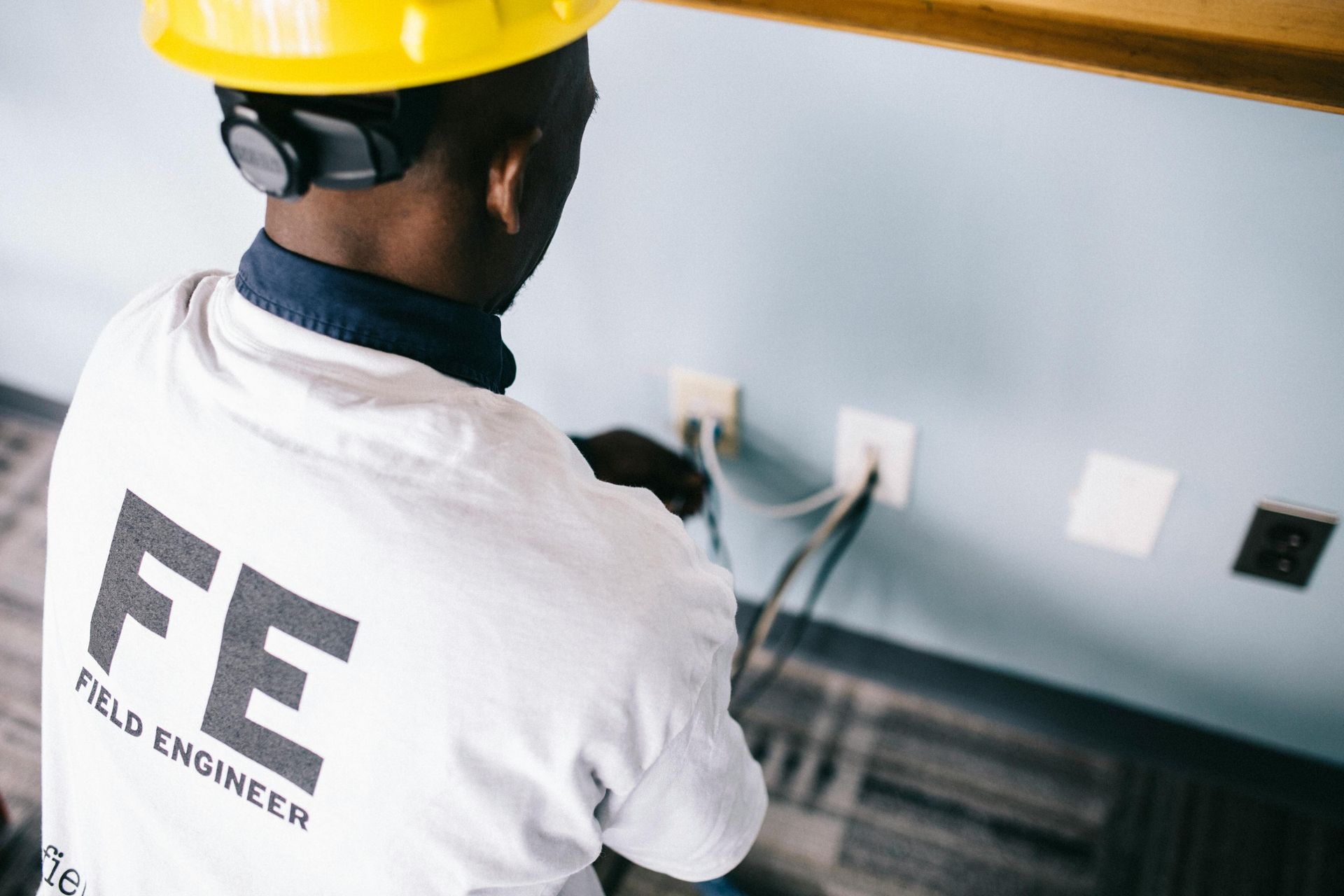 Field engineer working on electrical outlets; wearing yellow hard hat and white shirt with logo.