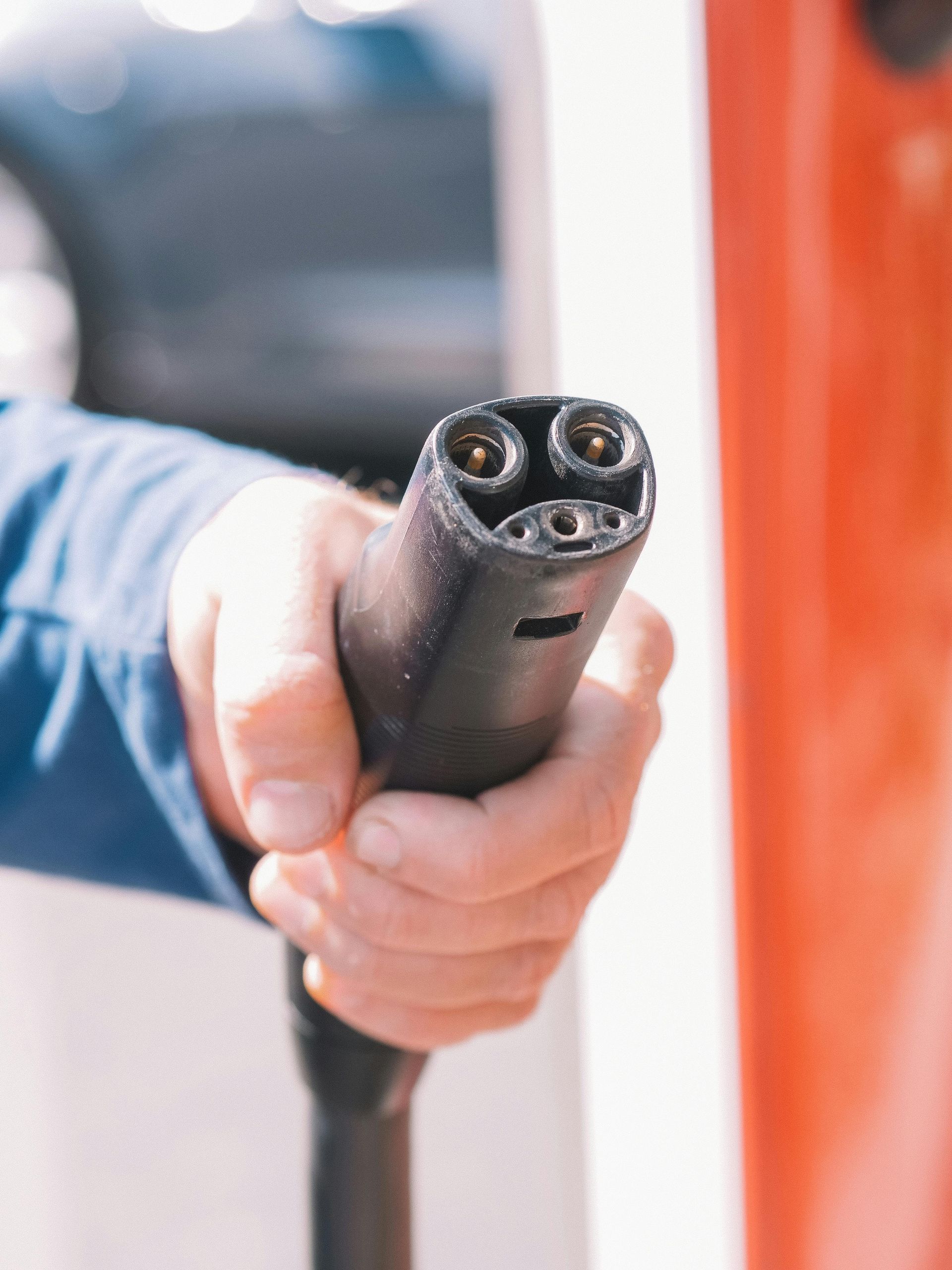 Hand holding a black electric vehicle charging connector, red and white charging station in background.