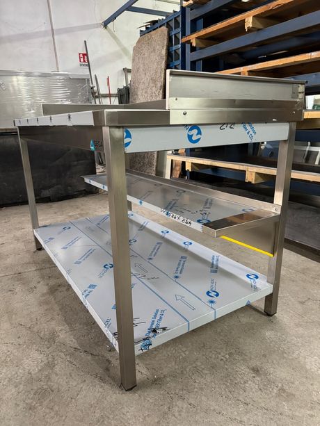 Tavolo da lavoro in acciaio inox con ripiano inferiore, all'interno di un magazzino industriale, ricoperto da pellicola protettiva.