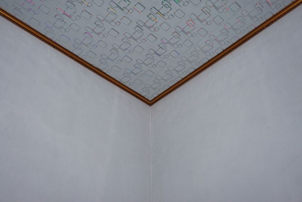 Corner of a room with white walls and patterned ceiling, framed with a brown border.