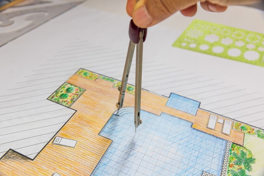 Hand using a compass on a colorful blueprint of a pool, plants, and patio.