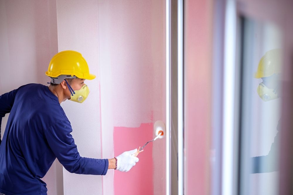 Painter in yellow hard hat and respirator painting a wall pink.