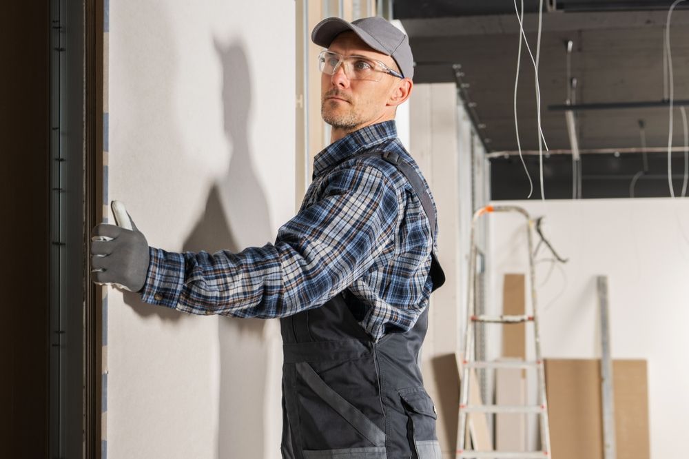 Construction worker inspecting drywall installation. Wearing work clothes, cap, and gloves. Indoor setting.