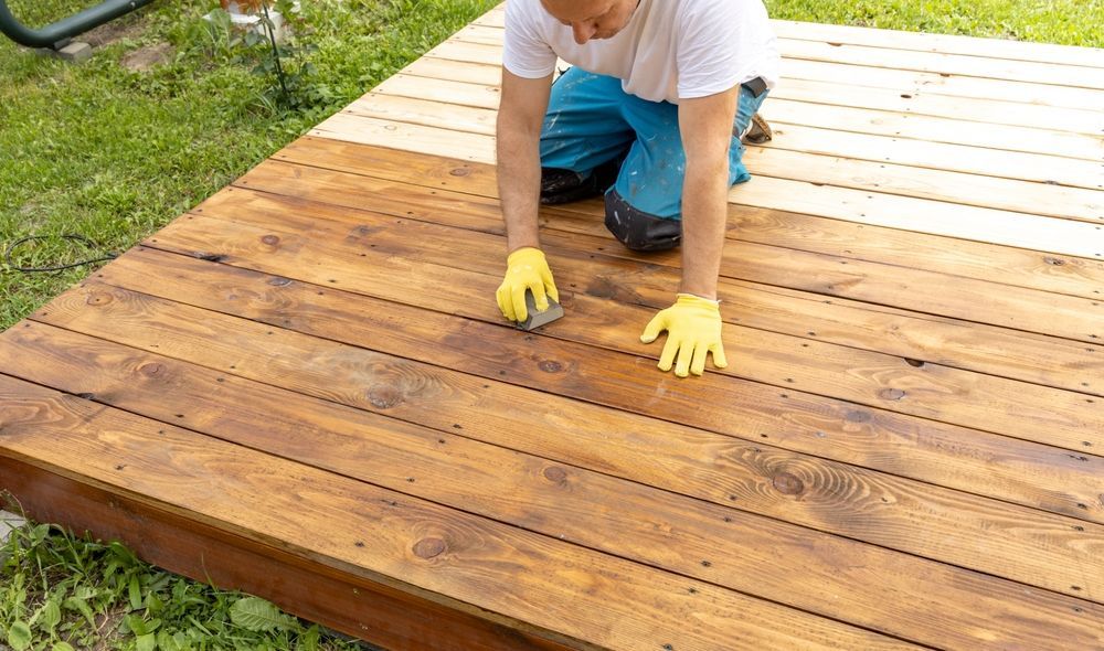 Person kneeling on wooden deck, applying stain with yellow-gloved hands.