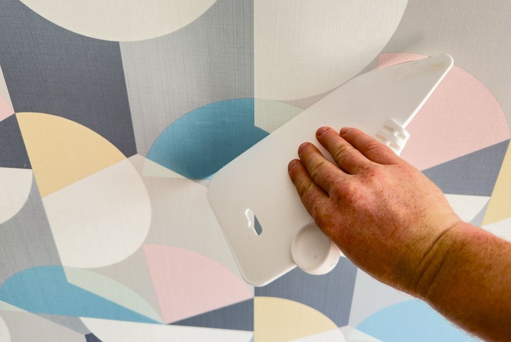 Hand using a smoothing tool on wallpaper with colorful geometric shapes.