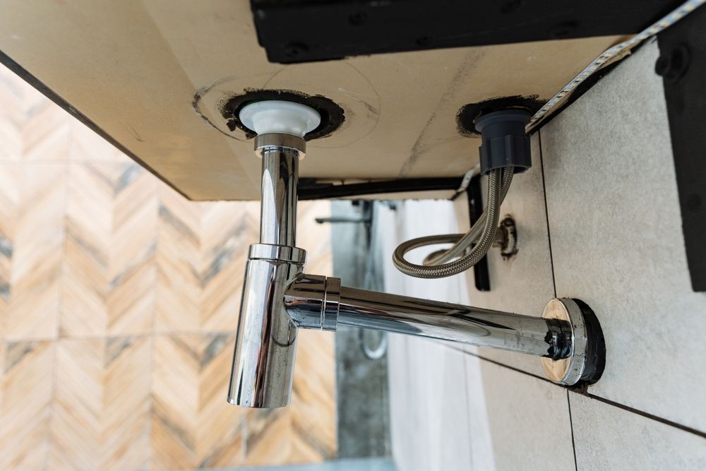 Chrome plumbing under a sink: drain pipe, water supply line, mounted on light wood and tiled wall.