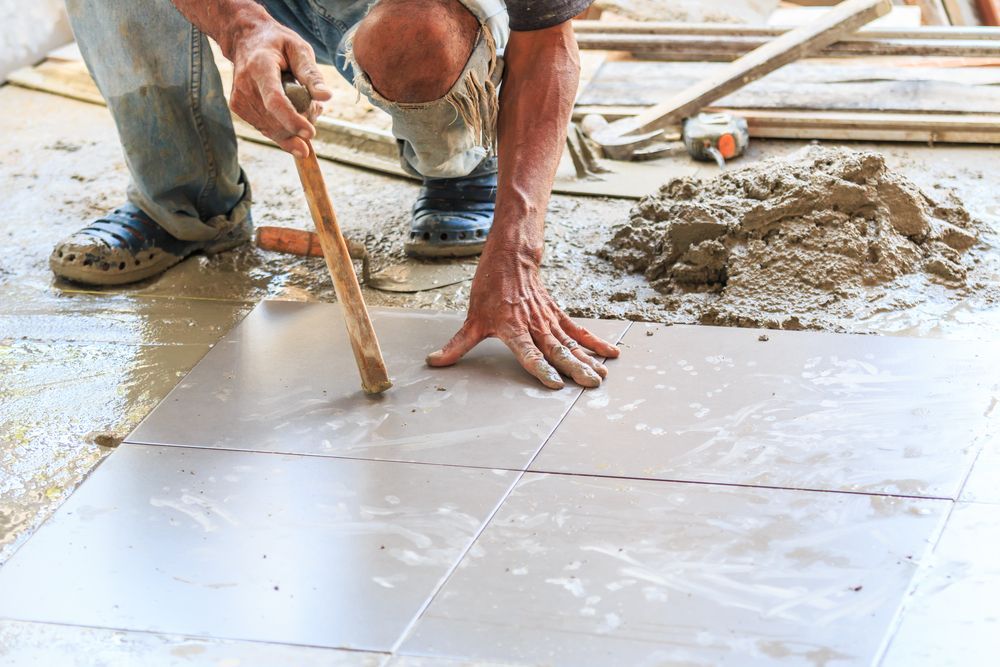 A person is laying floor tiles with a trowel on a construction site.