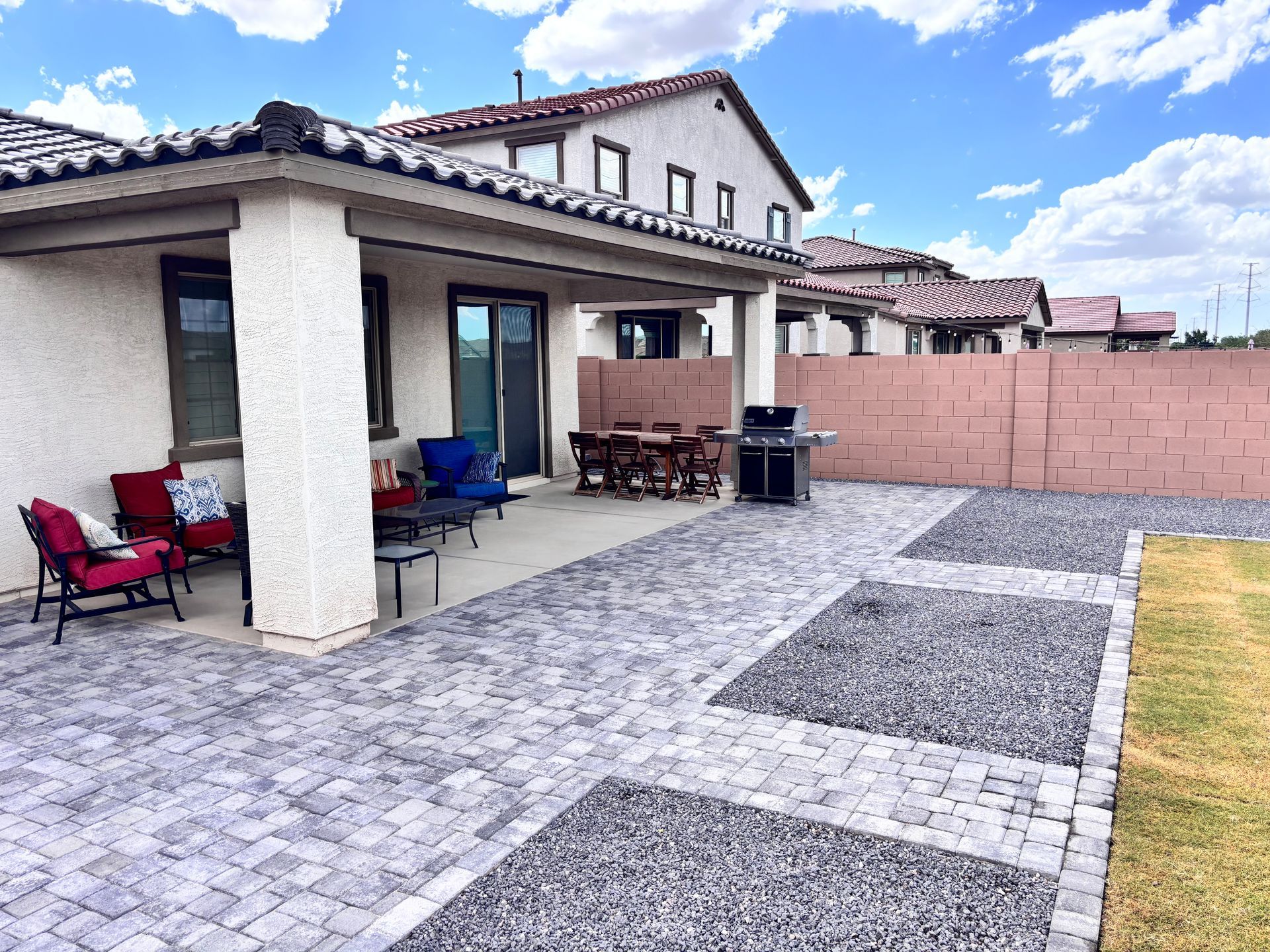 Backyard patio with seating, grill, and paved walkways. Beige house and red brick wall under a blue sky.