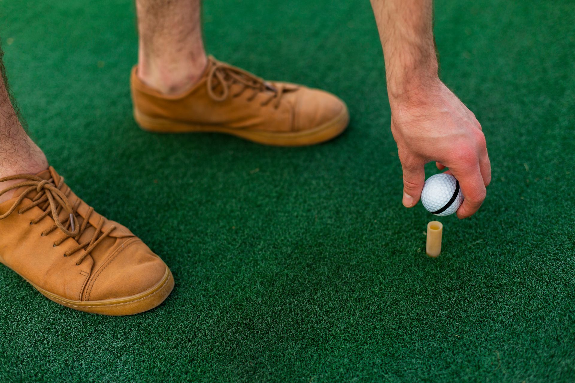 A person placing a small ball onto a wooden peg on a green surface, wearing brown shoes.