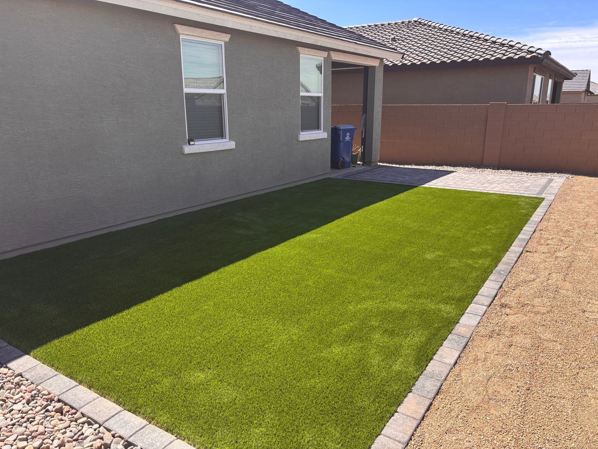 Backyard with green turf, bordered by paving stones and gravel, beside a stucco home.
