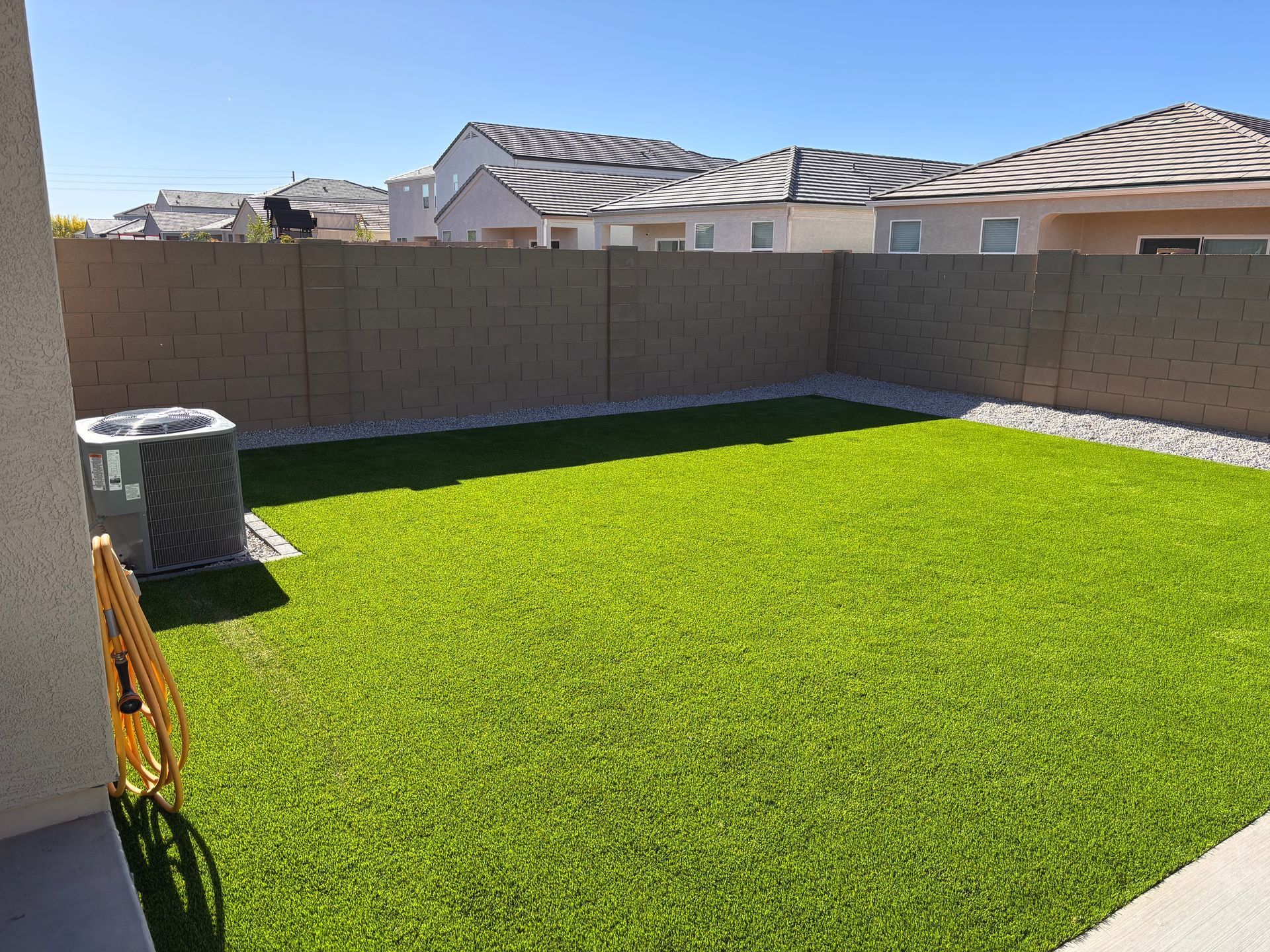 Backyard with artificial green grass, stone border, and concrete wall. An AC unit is on the side.