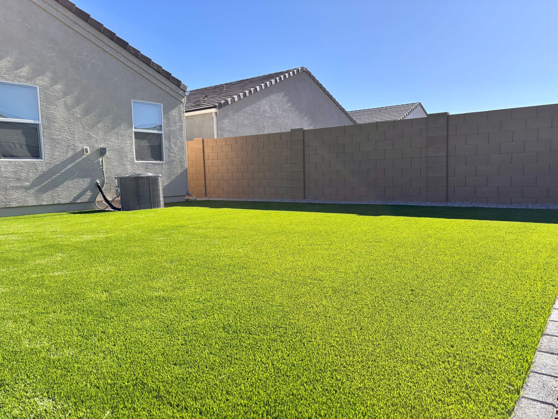 Backyard with green grass, tan wall, and white stucco house under a blue sky.