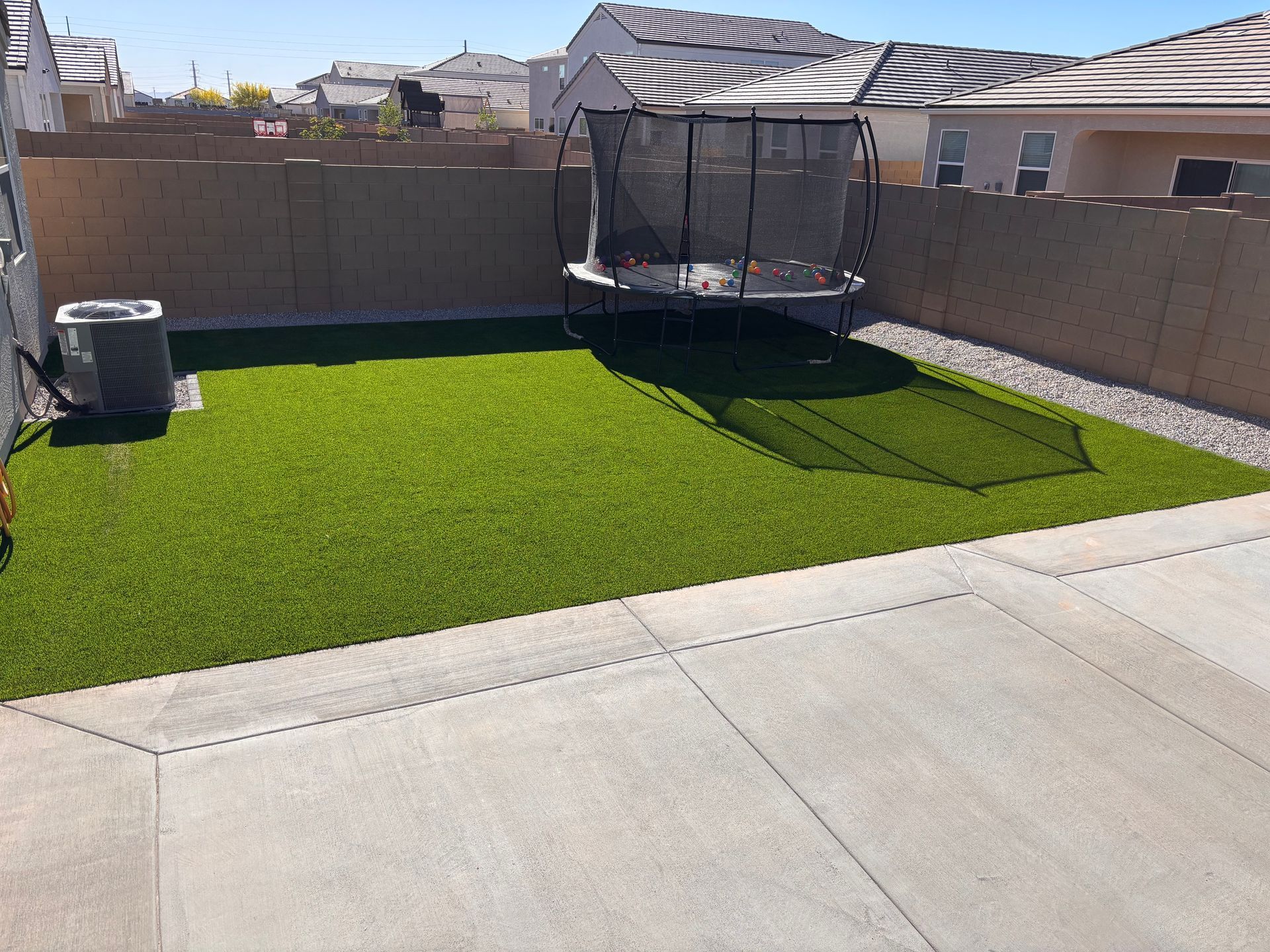 Backyard with green turf, trampoline, air conditioner, and concrete patio.