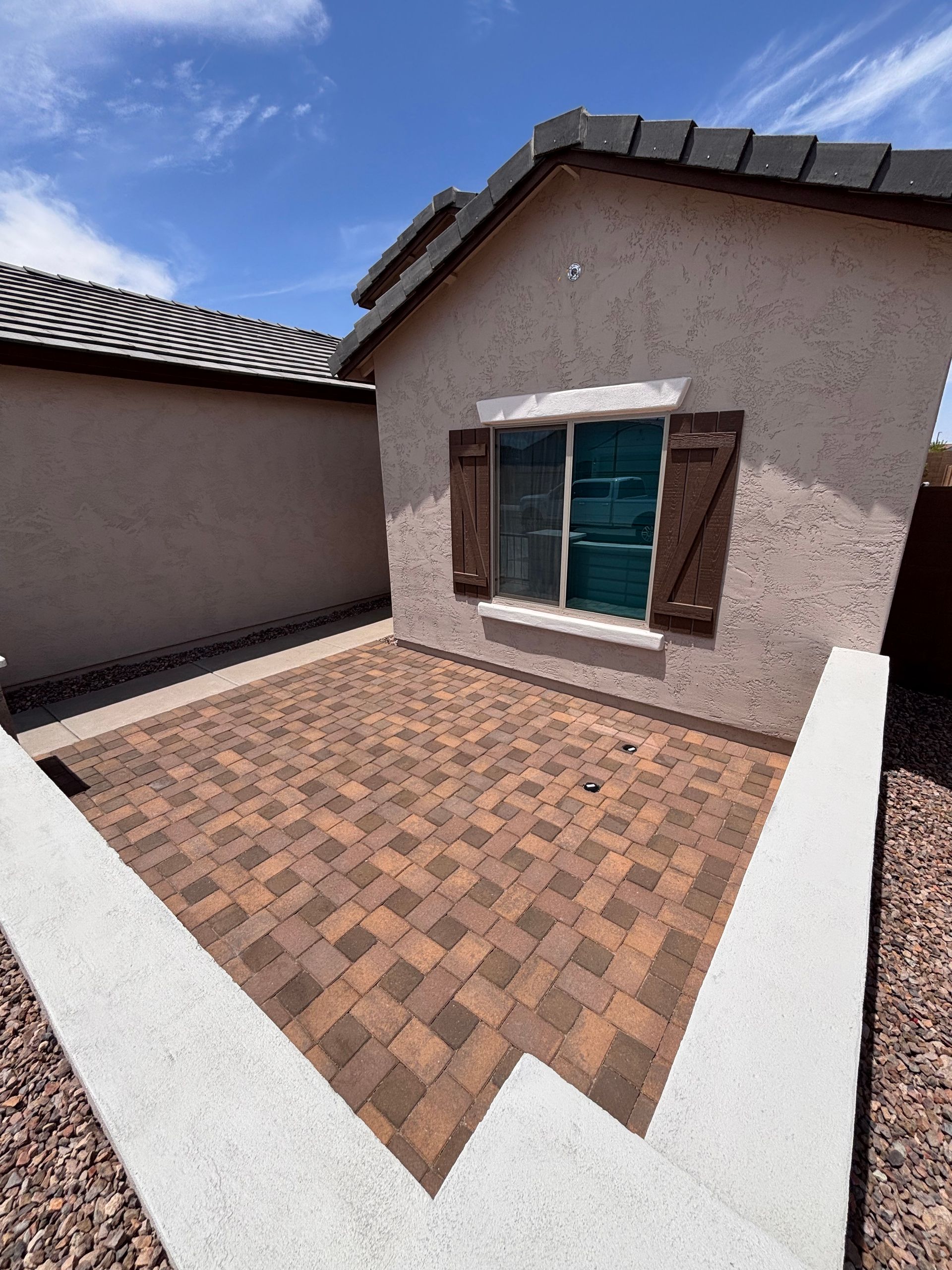 Brick patio with white retaining walls, tan stucco building, wooden shutters, and blue sky.