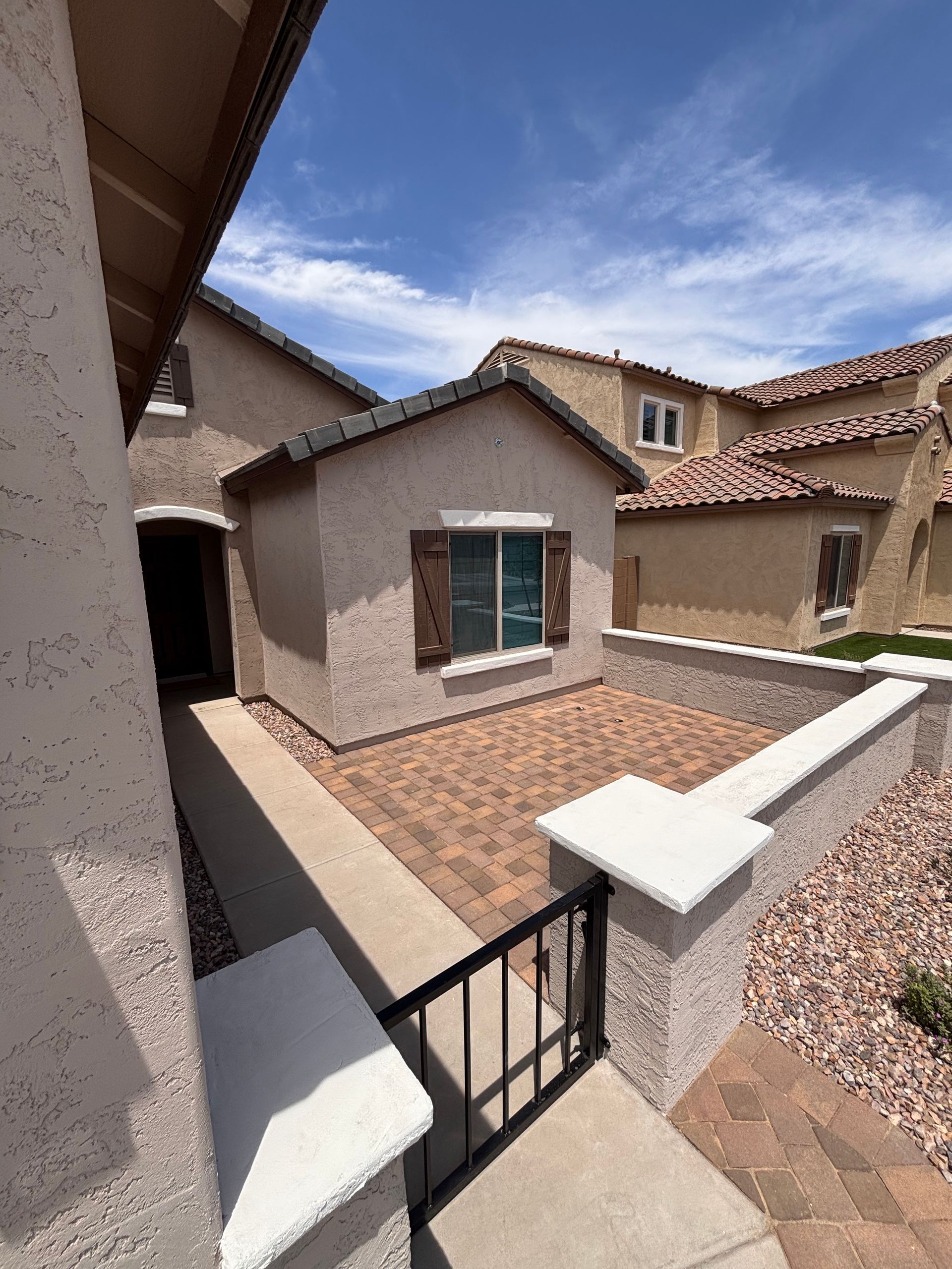 Exterior view of a small stucco building with a brick patio and a black gate under a blue sky.