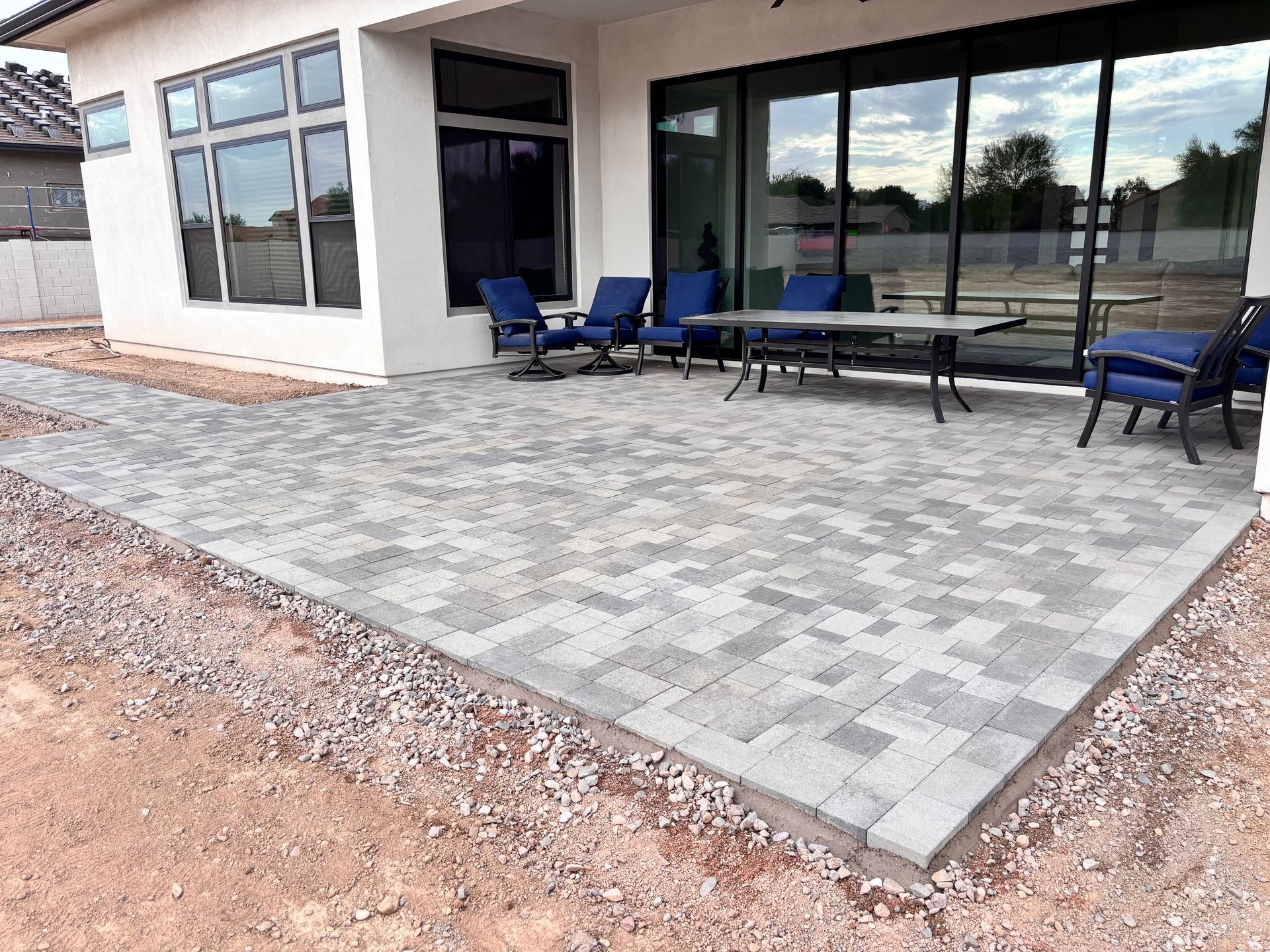 Backyard patio with gray brick pavers, blue chairs, and a table, adjacent to a white house with large windows.