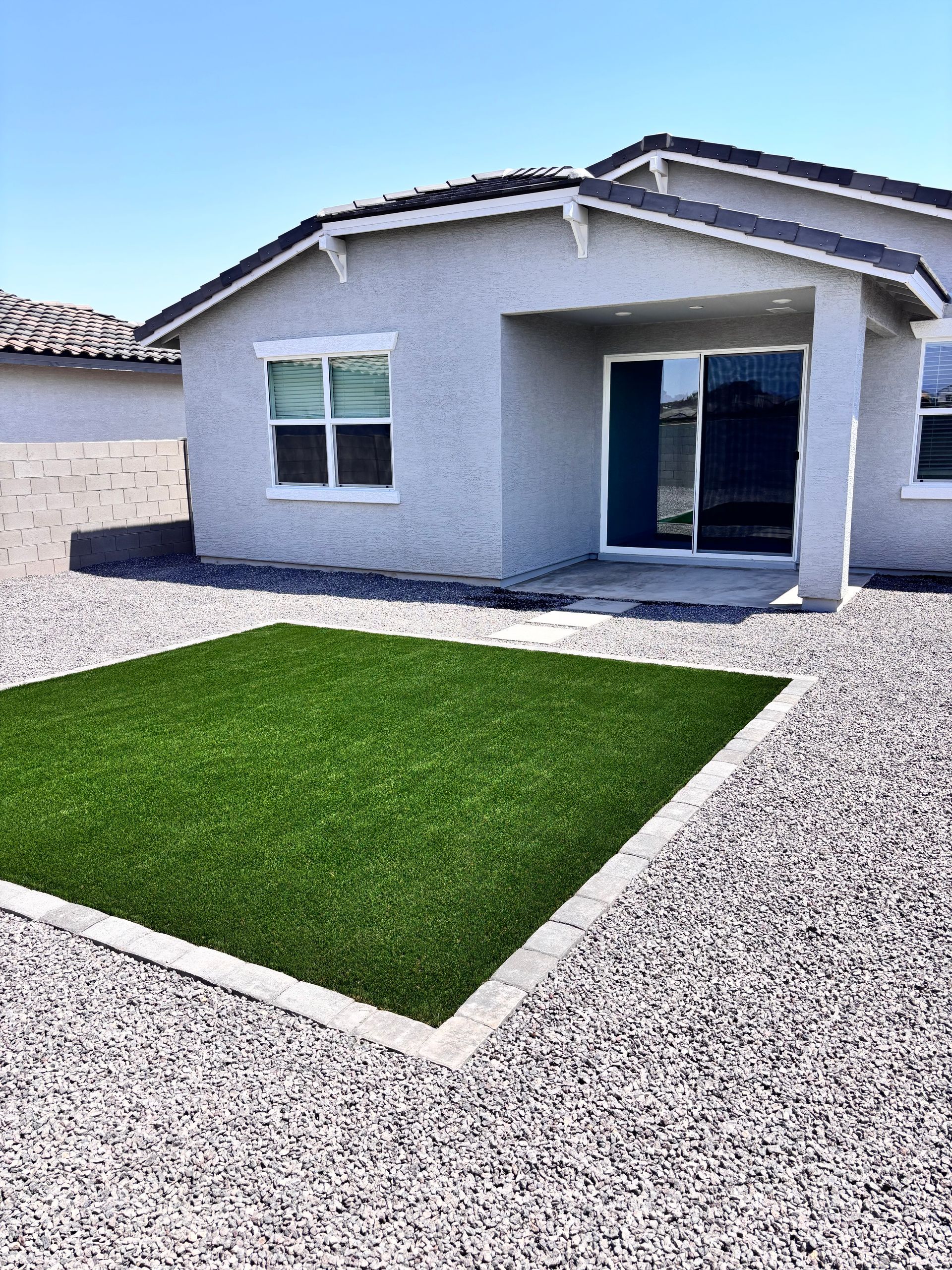 Backyard with a square of green artificial turf, surrounded by gravel, and a light gray house.