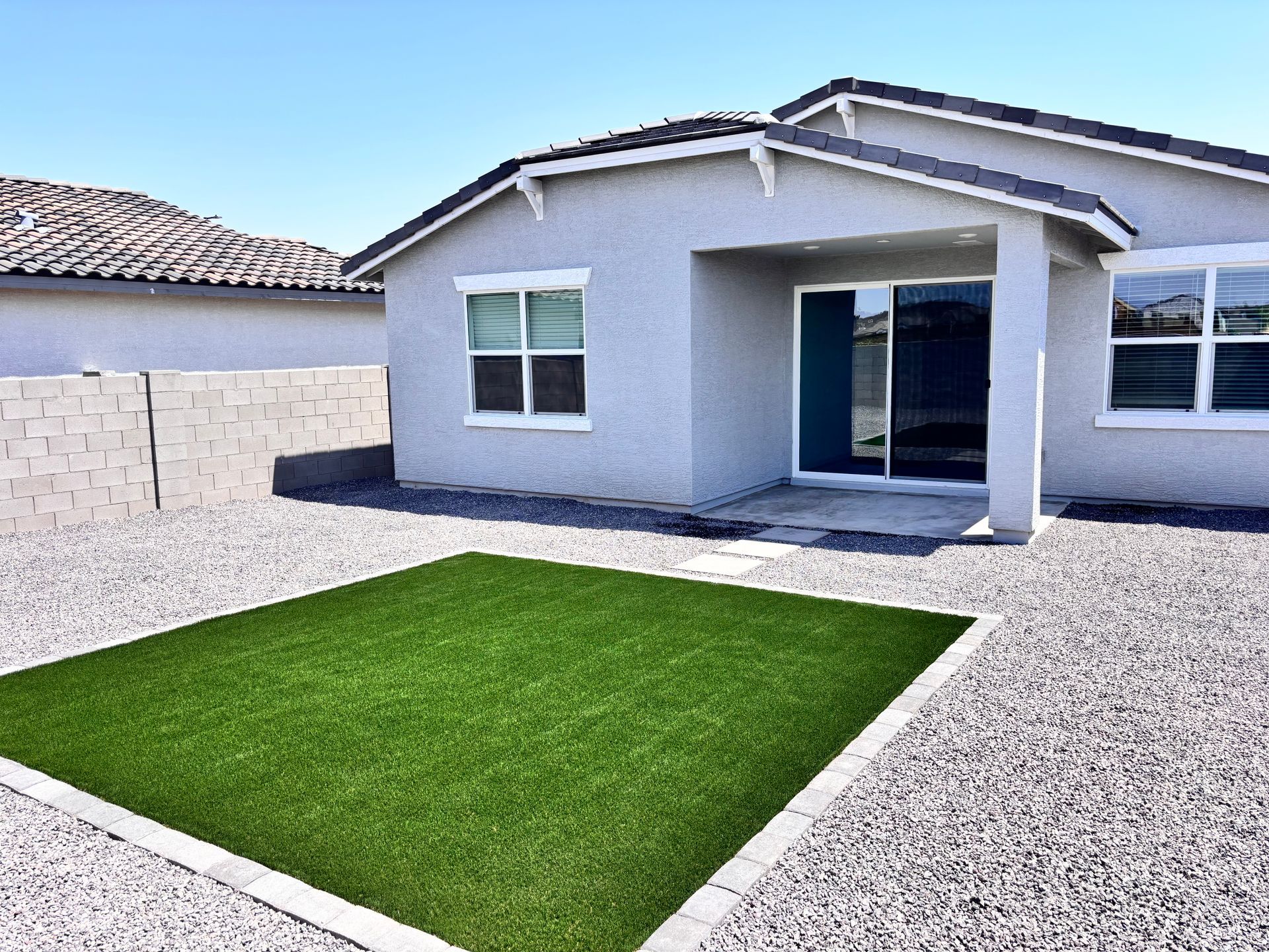 Backyard with artificial grass rectangle surrounded by gravel, gray house and walls under blue sky.