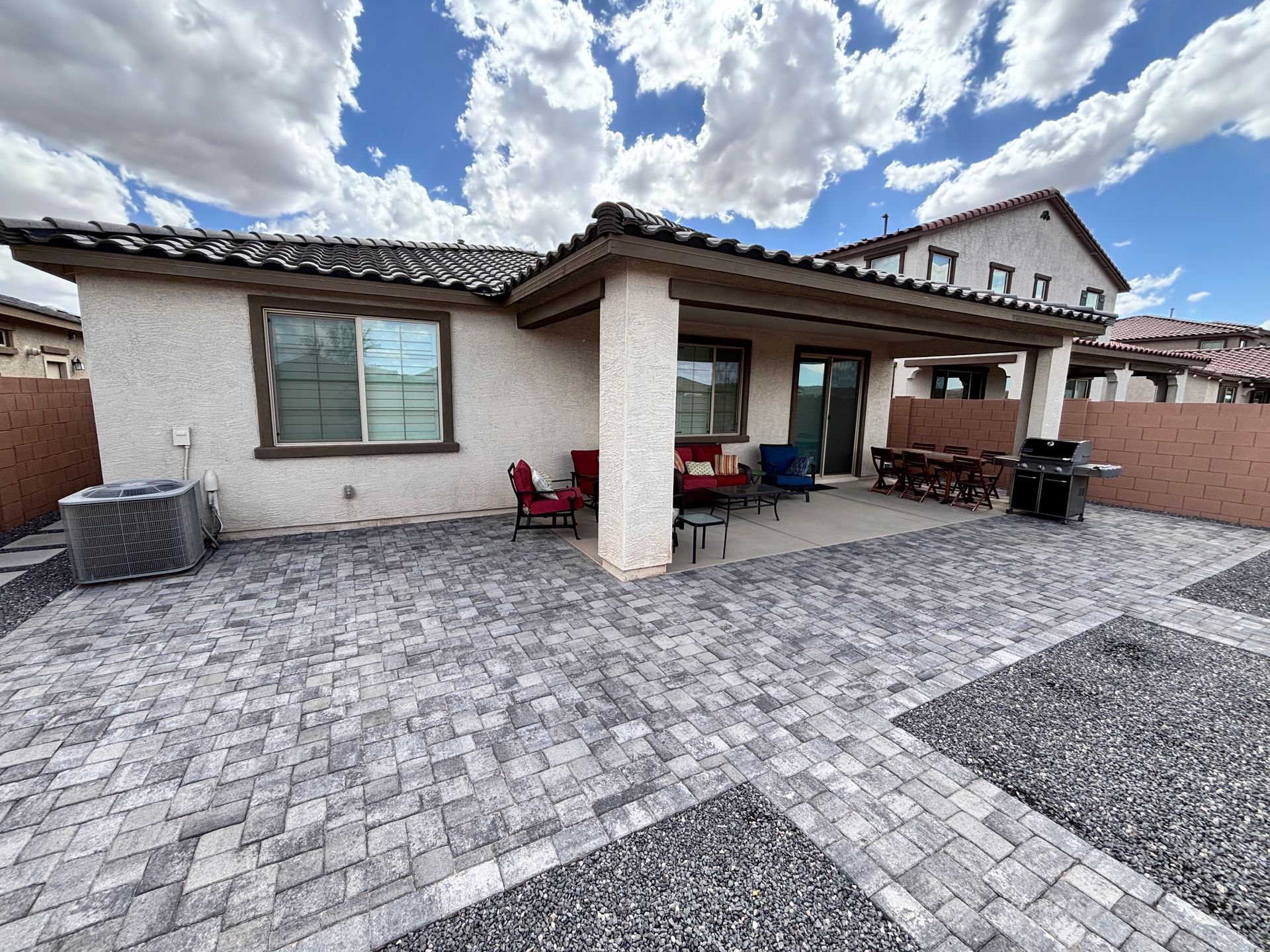 Backyard with patio, pavers, red chairs, and grill; light stucco house under a cloudy sky.