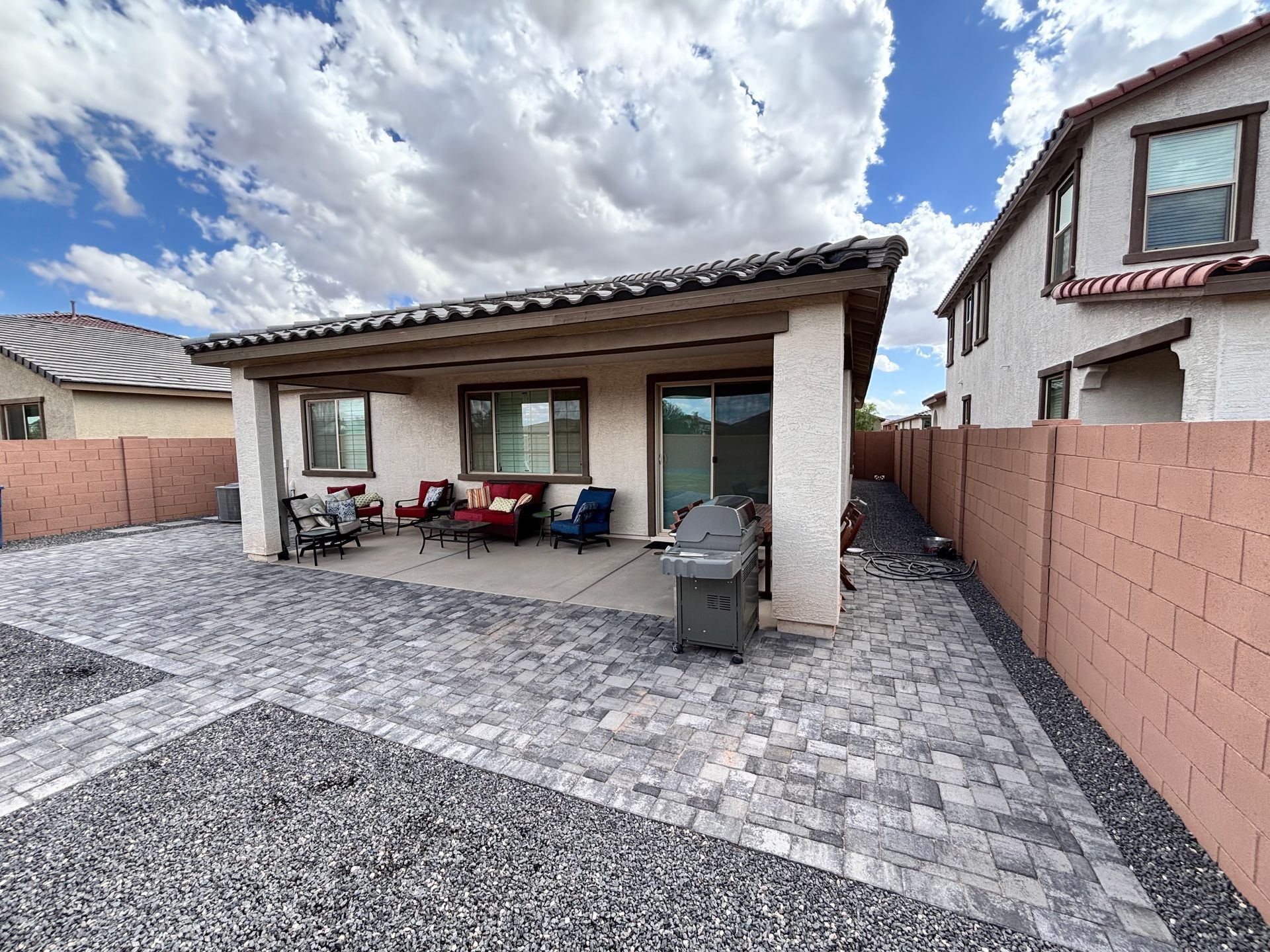 Backyard patio with gray pavers, brick walls, and a blue sky.  A grill, chairs, and a sliding glass door are visible.