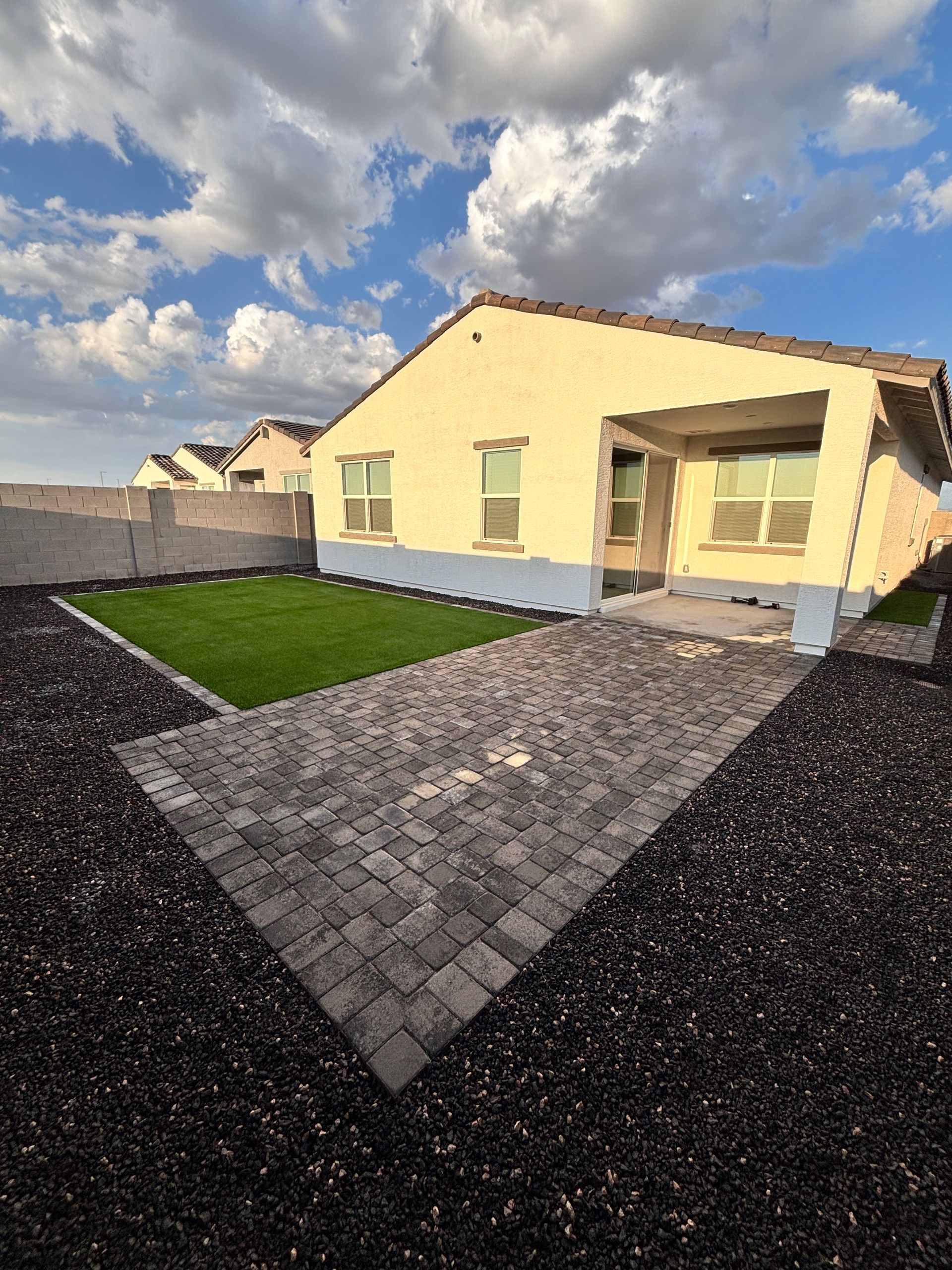 Beige house with gray paver walkway and green grass, set against a blue sky with clouds.