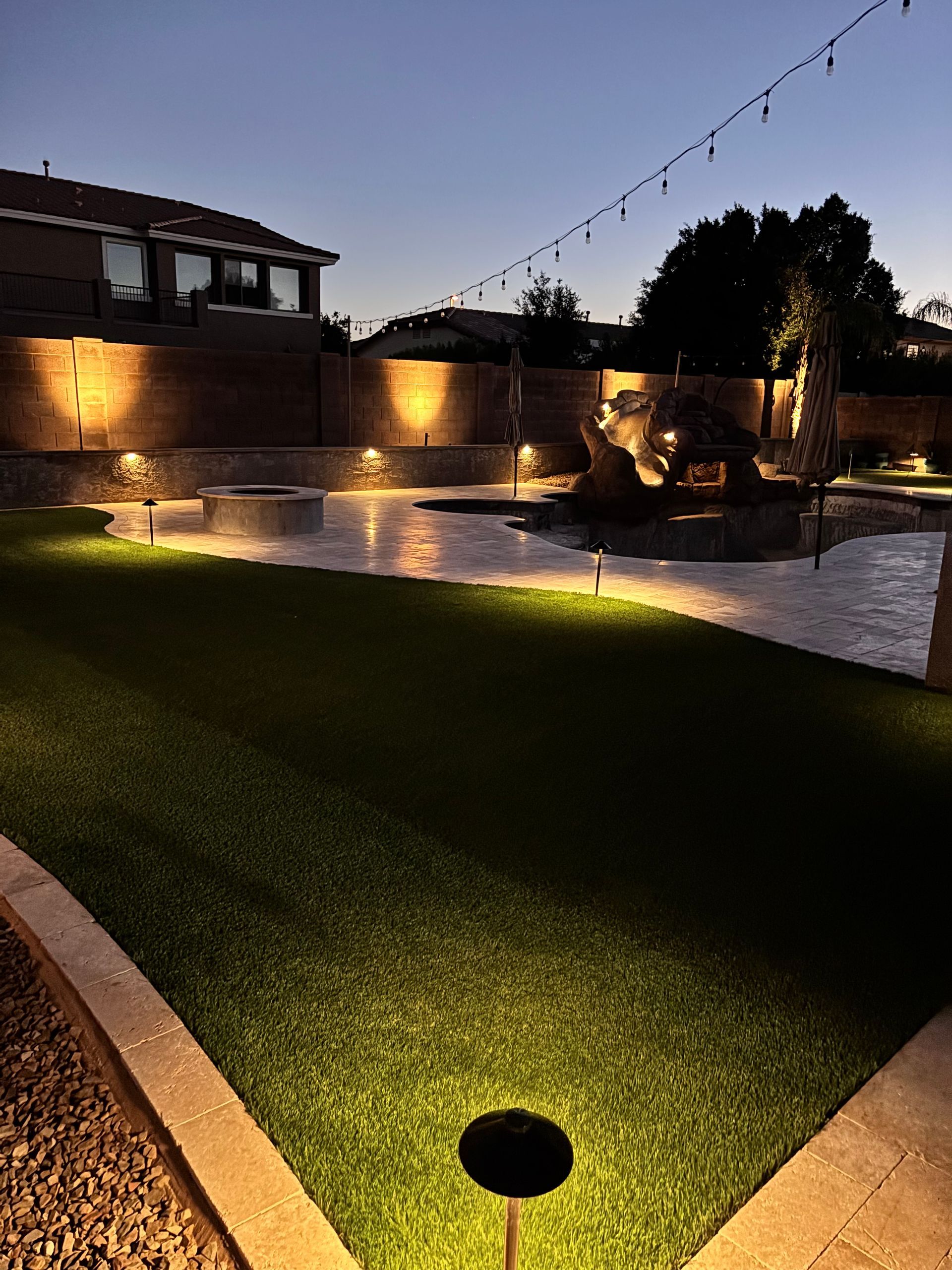 Backyard at dusk with artificial turf, patio, and landscape lighting illuminating the scene.