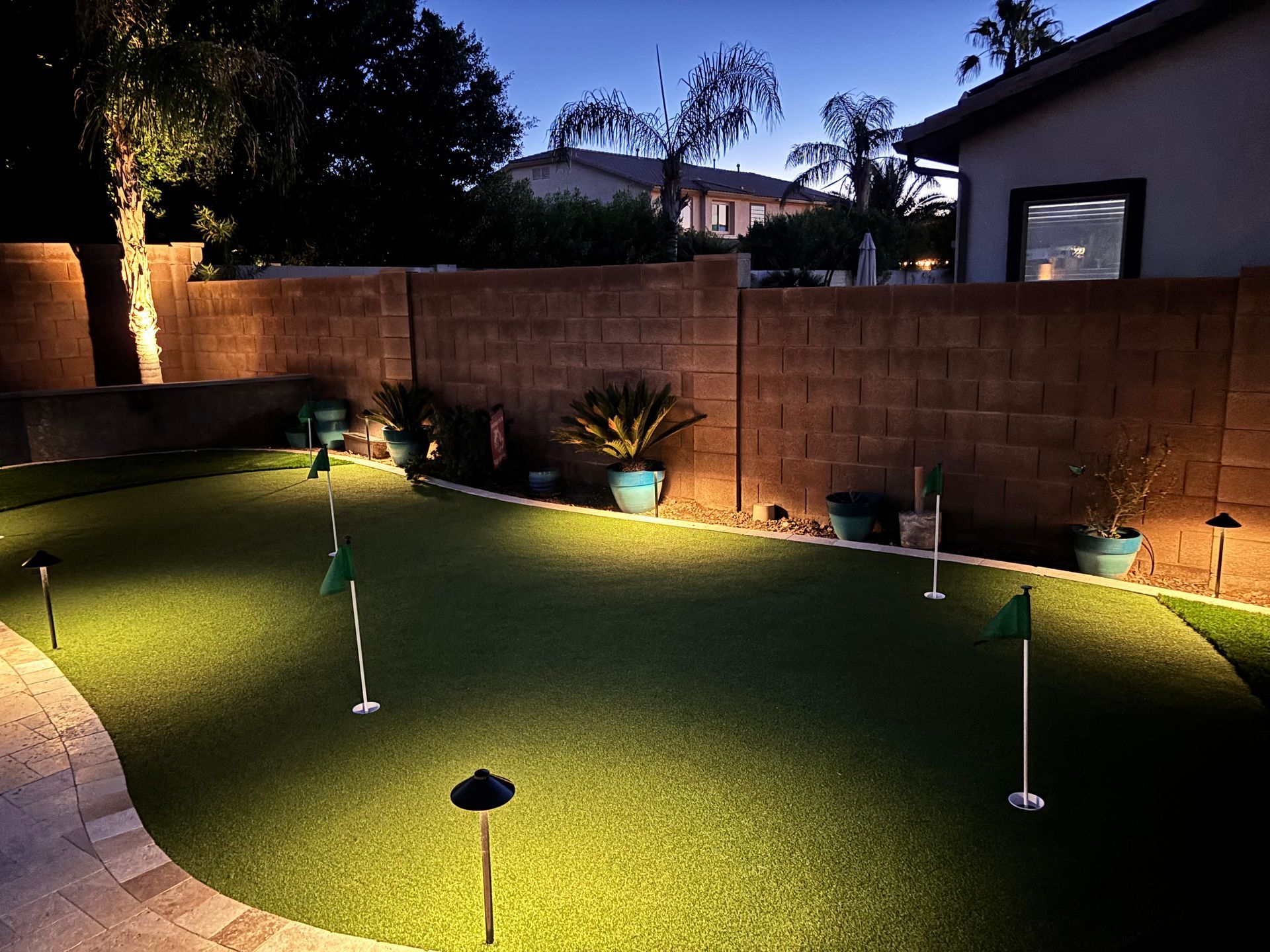 A backyard putting green with spotlights, flags, and a brick wall, at dusk.