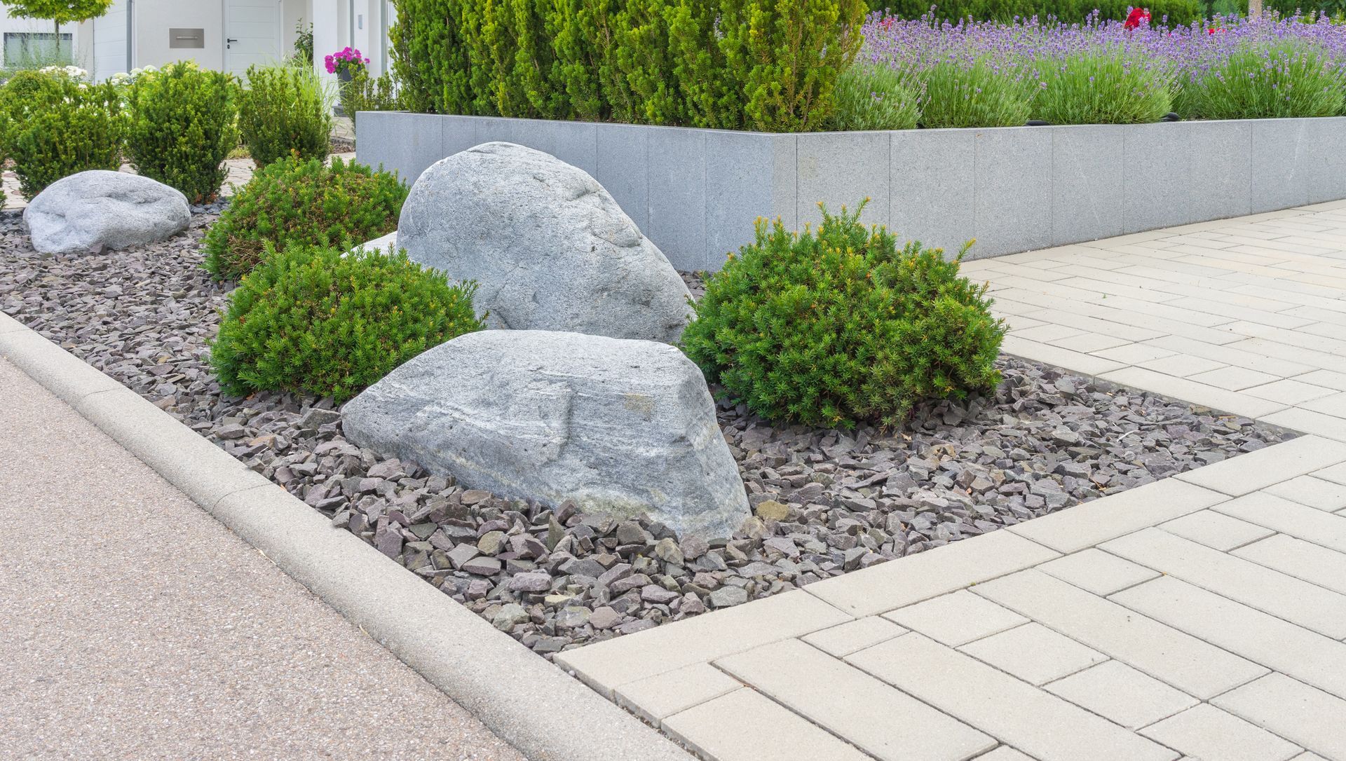 Garden bed with large rocks, green shrubs, and bark mulch next to a paved pathway.