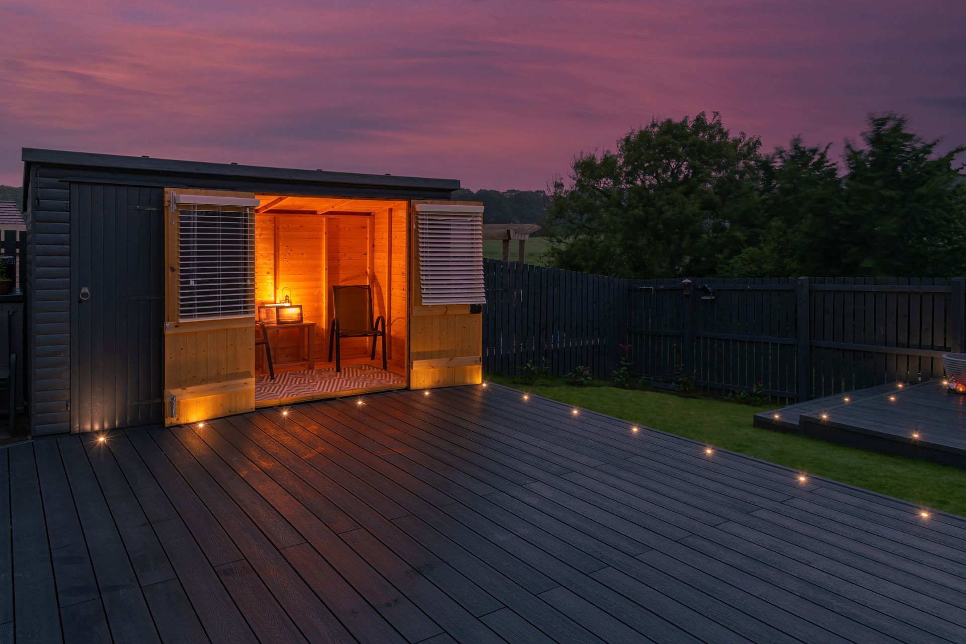 Shed with open doors and warm light on a wooden deck at dusk, surrounded by a dark fence and grass.