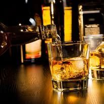 Whiskey being poured into a glass, bottles in the background, on a wood surface.