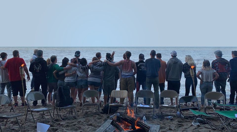 Group stands on beach, arms around each other, facing ocean. A fire burns in front, chairs are set up.
