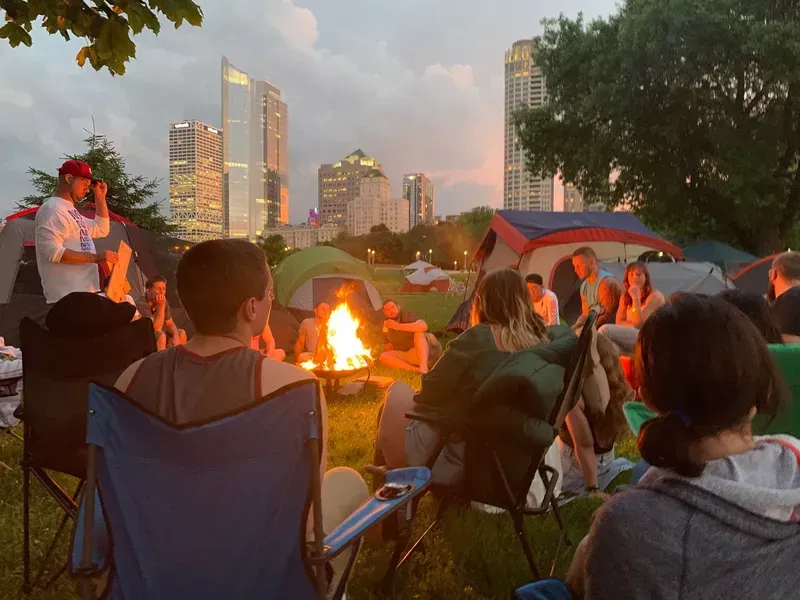 Campfire gathering in a park, tents, people in chairs, city skyline in background, sunset colors.
