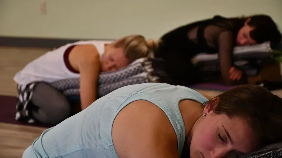 People resting on yoga mats, supported by blankets. Indoors, dim lighting.