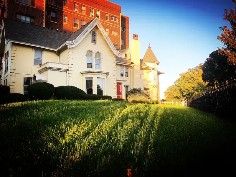 Cream-colored Victorian house with a red door, set against a lawn, and a tall building in the background under a sunny sky.